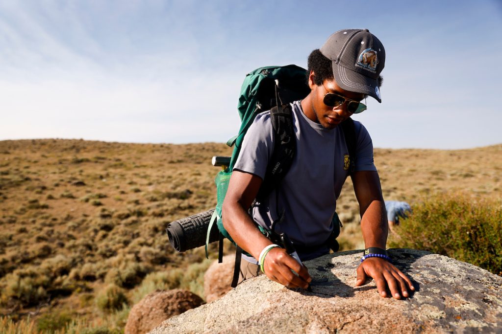 Young Black man wearing cap studies rock while conducting field research on Wyoming landscape.