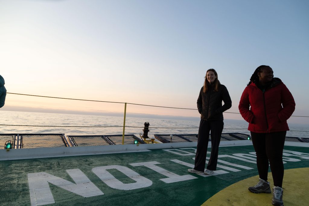 A woman in a dark coat and pants stands on the deck of a boat, with a view of the ocean in the background.