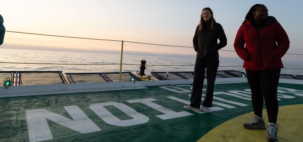 A woman in a dark coat and pants stands on the deck of a boat, with a view of the ocean in the background.